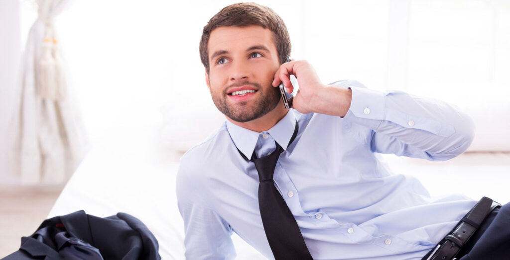 Mann in Businessoutfit liegt auf Hotelbett mit gelockerter Krawatte und telefoniert mit dem Smartphone, links daneben liegt sein Jacket auf dem Bett