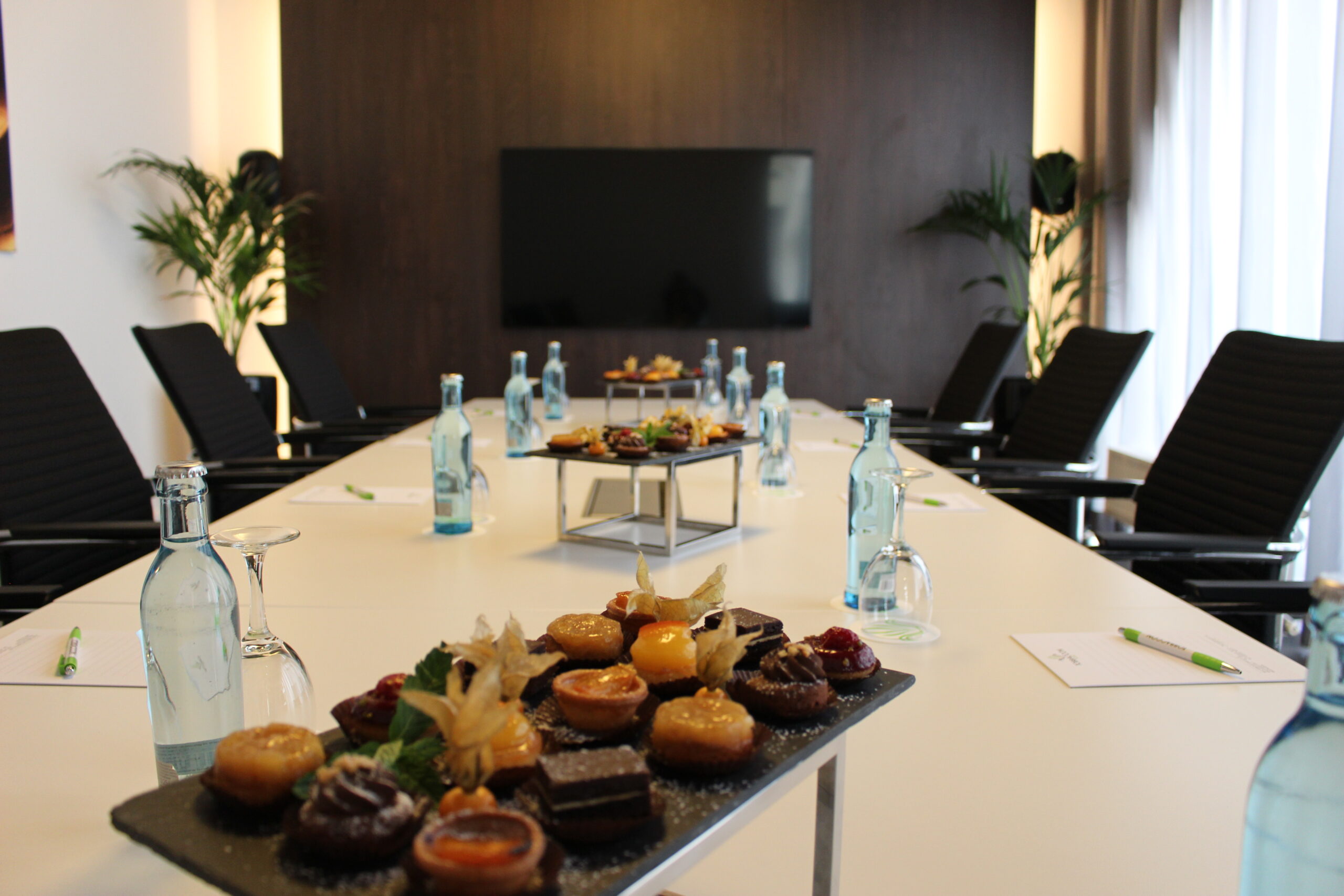 Blick über den Tisch in den Tagungsraum Mojito, links und rechts dunkle Stühle, auf dem Tisch blaue Wasserflaschen, Wassergläser, Blöcke mit Kugelschreibern und stilvoll dekorierte süße Snacks; hinten braune Wand mit Flachbildschirmfernseher und links und rechts davon Grünpflanzen