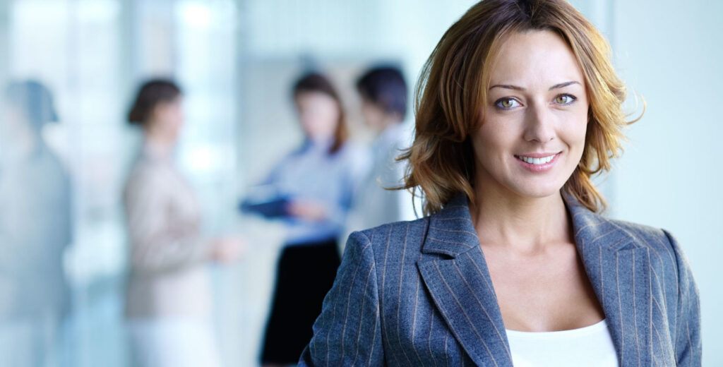 Lächelnde junge Frau in Businesskleidung, im Hintergrund weitere Personen in Businessoutfits