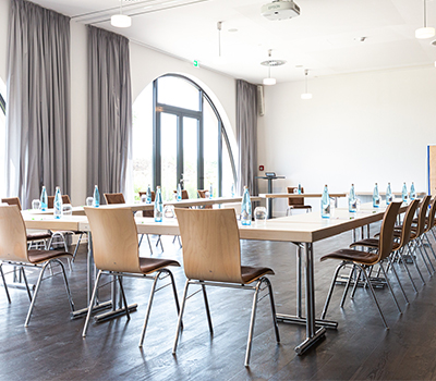 Lichtdurchfluteter, eingedeckter Tagungsraum des Hotels Tobbaccon, hellbraune Tische und Stühle im Quadrat gestellt, links bodentiefes halbrundes Fenster und hellgraue Vorhangschals