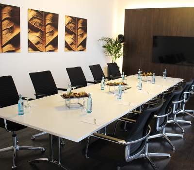 Kleiner, eingedeckter Tagungsraum des Hotels Tobbaccon mit großem weißen Tisch (darauf blaue Wasserflaschen, Wassergläser, Block und Kugelschreiber an jedem Platz sowie Snacks), schwarze Stühle am Tisch, links an der Wand drei Bilder von Tabakblättern, in der Ecke eine Grünpflanze, an der hinteren dunkelbraunen Wand Flachbildschirmfernseher