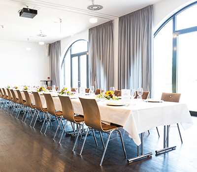Festlich gedeckte Tafel in lichtdurchflutetem Veranstaltungsraum des Hotels Tobbaccon, rechts bodentiefe halbrunde Fenster und hellgraue Vorhänge