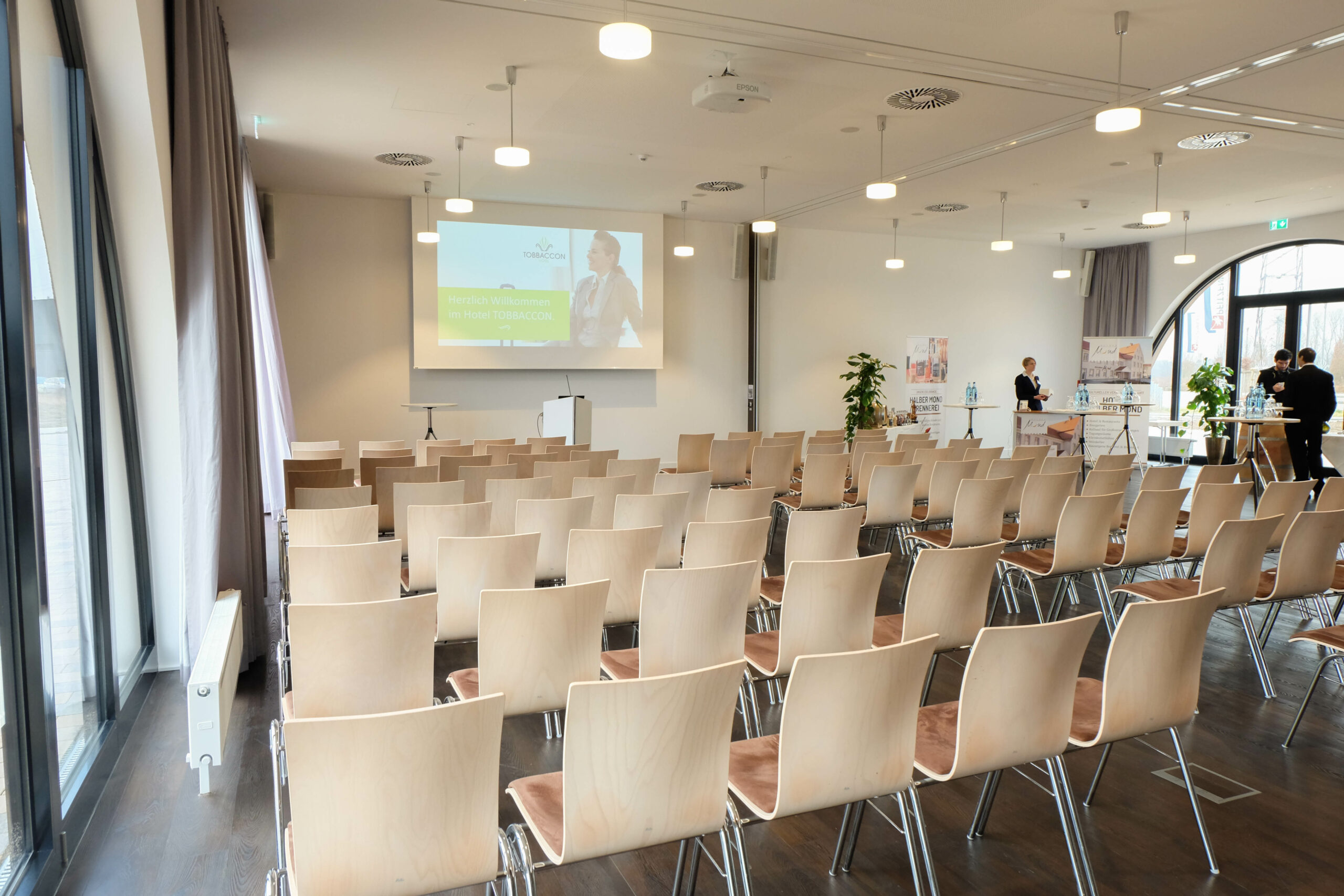 Blick in den Tagungsraum Cuba Libre des Hotels Tobbaccon, der mit mehreren Reihen heller Stühle bestuhlt ist, links Fenster mit grauen Vorhangschals, vorne Leinwand, rechts halbrundes bodentiefes Fenster und Menschen an einem Werbestand