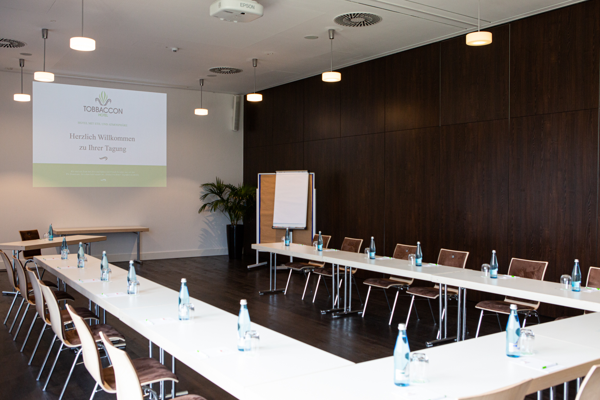 Blick in den Tagungsraum Cuba oder Libre mit eingedeckten Tischen in U-Form, vorne Leinwand mit Logo des Hotels Tobbaccon, auf der steht: „Hotel mit Stil und Atmosphäre, Herzlich willkommen zu Ihrer Tagung“, rechts Grünpflanze und Flipchart vor dunkelbrauner Wand, an der Decke kleine helle Hängelampen