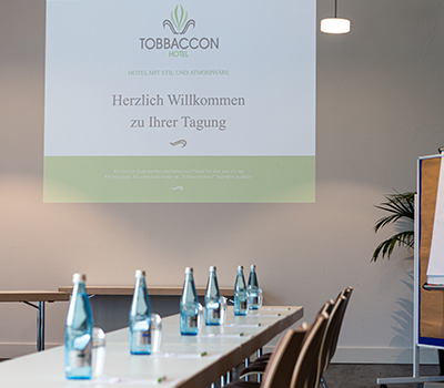 Blick über eingedeckten Tisch in Tagungsraum des Hotels Tobbaccon mit Blick auf Leinwand mit Logo des Hotels Tobbaccon, auf der steht: „Hotel mit Stil und Atmosphäre, Herzlich willkommen zu Ihrer Tagung“; rechts Flipchart und Grünpflanze